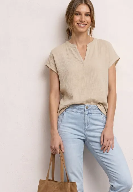 Een vrouw draagt een beige top met korte mouwen, lichtblauwe spijkerbroek en houdt een lichtbruine tas vast.