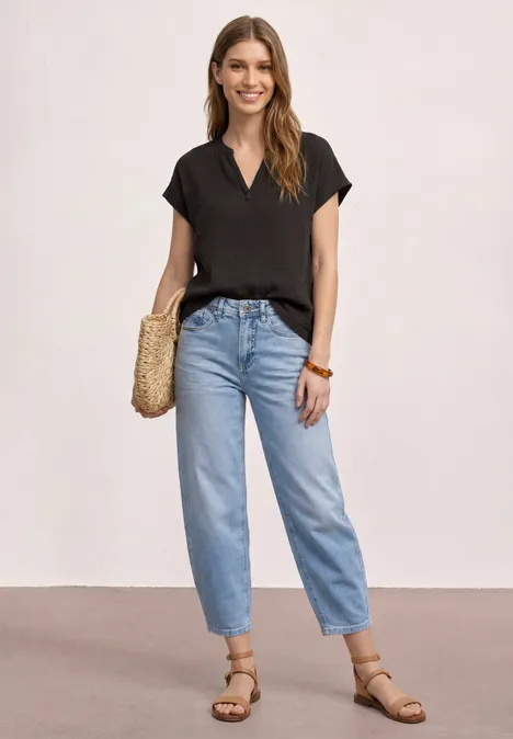 Femme en t-shirt noir, jean mom bleu clair, sandales plates et sac en paille sur fond uni.