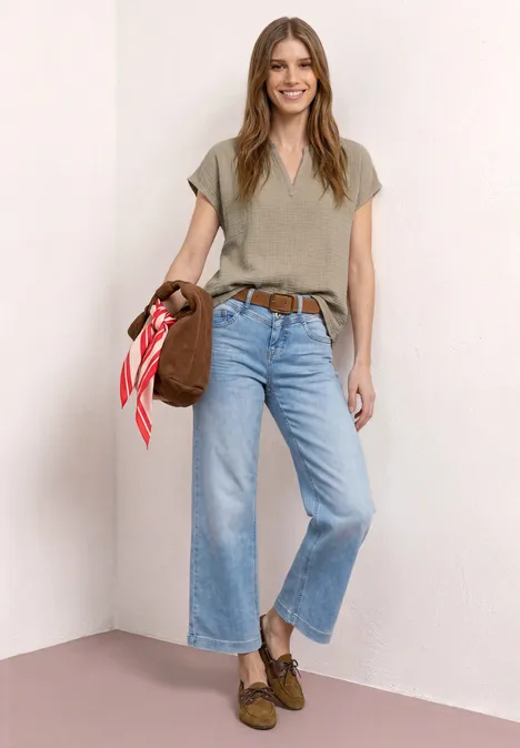 Een vrouw staat in een hoek, draagt een kaki V-hals shirt, lichtblauwe wijde cropped jeans, bruine loafers en een tas met rood sjaaltje.