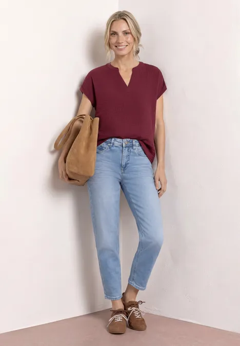 Frau mit weinrotem Kurzarmshirt, hellblauen Jeans, braunen Schnürschuhen und großer Wildledertasche vor hellem Hintergrund.