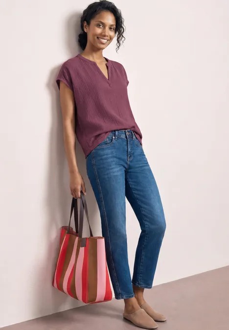 Femme en haut violet, jean bleu et sac rayé coloré devant un fond clair.