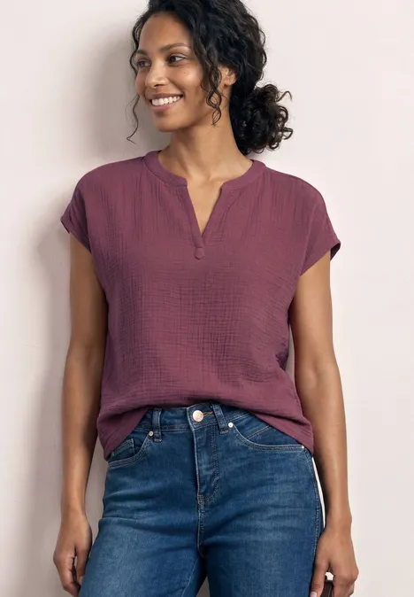 Femme porte un top bordeaux à manches courtes et un jean bleu, posant devant un fond clair.