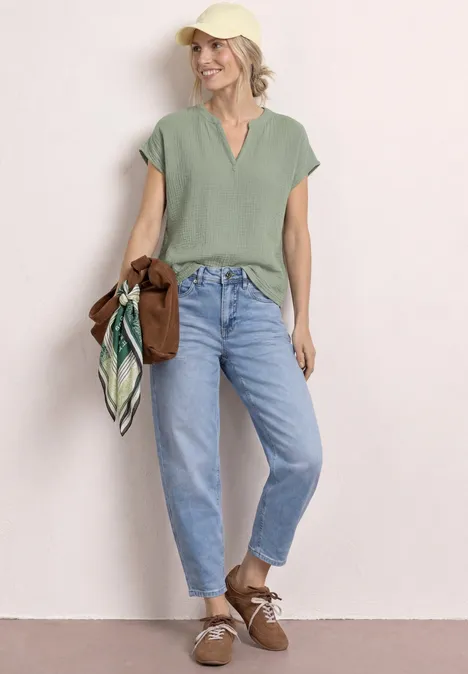Vrouw in casual outfit: groen T-shirt, wijde mom-jeans, bruine schoenen, ze houdt een trui en sjaal vast.