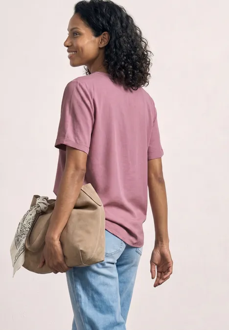 Person von der Seite in einem pastellrosa T-Shirt und blauen Jeans, die eine beige Tasche hält.