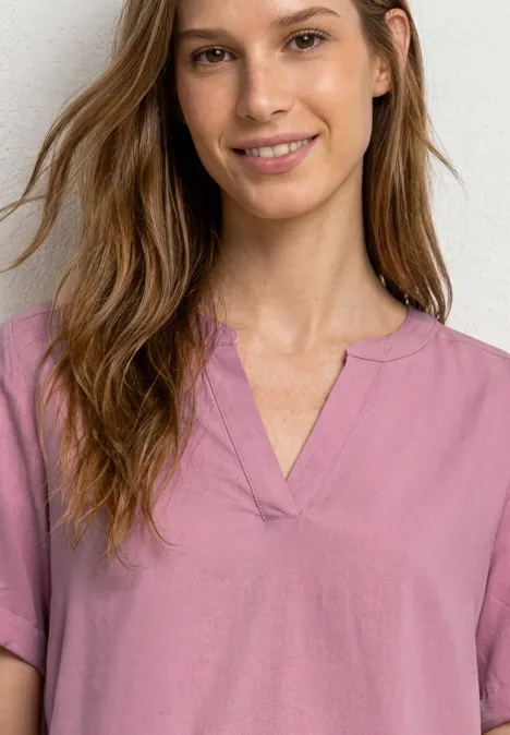 Blouse met gespleten hals en omslag clouded blush Vrouw met onscherp gezicht, golvend bruin haar en roze kort mouwtoppie met V-hals, neutrale achtergrond.