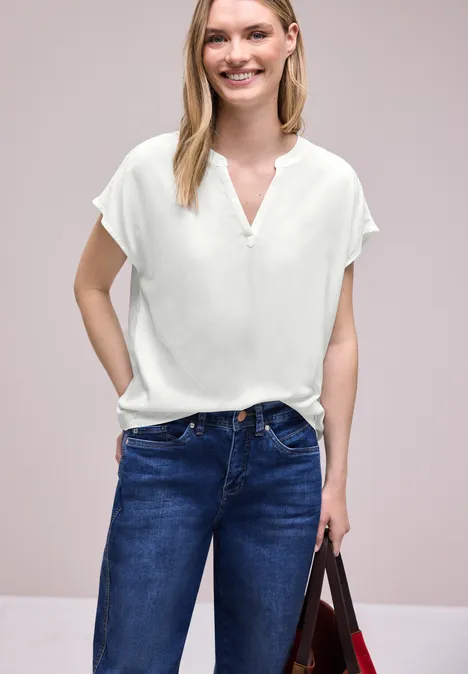 Vrouw in witte blouse met V-hals en blauwe jeans, met tas voor een neutrale achtergrond.