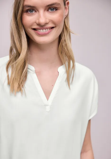 Een persoon draagt een witte blouse met V-hals tegen een neutrale achtergrond.