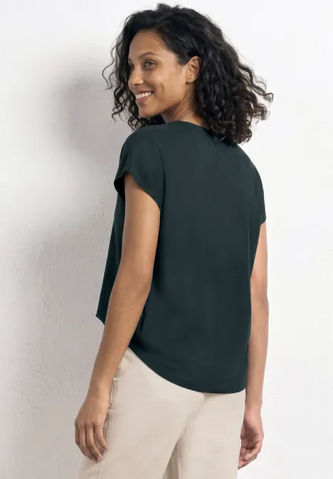 Eine Frau von hinten in einer locker sitzenden, dunkelgrünen Bluse und hellen Hosen.