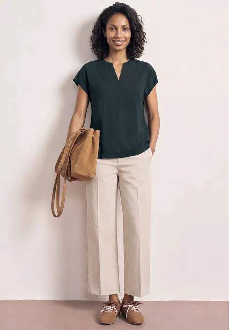 Frau in schwarzer Bluse, beigen weiten Hosen, braunen Schuhen und Tasche vor neutralem Hintergrund.