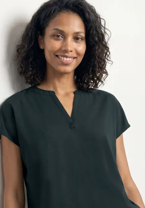 Frau mit lockigem Haar trägt schlichte, dunkelgrüne Kurzarmbluse vor weißem Hintergrund.