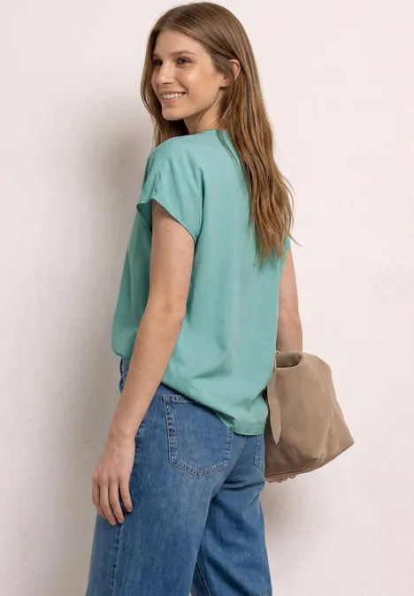 Femme en t-shirt turquoise, jean bleu et sac beige, regard par-dessus l'épaule.