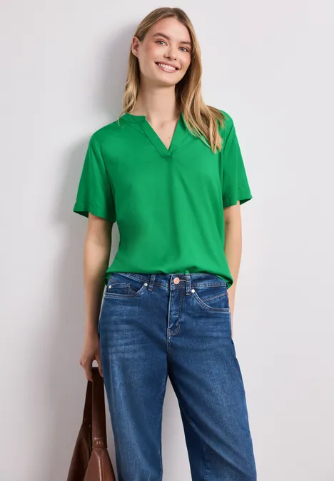 Frau in grüner V-Ausschnitt-Bluse, blauen Jeans und brauner Tasche vor neutralem Hintergrund.