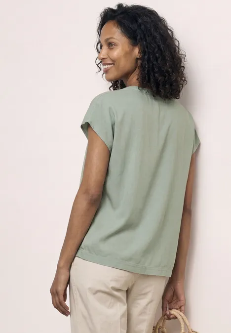 Frau von hinten in locker sitzender grüner Bluse und hellen Hosen vor neutralem Hintergrund.