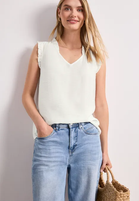 Frau trägt weißes ärmelloses V-Shirt, helle Jeans und hält eine geflochtene Strohtasche.