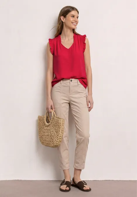Vrouw in rode volantblouse, beige broek en sandalen met een strotas voor een lichte muur