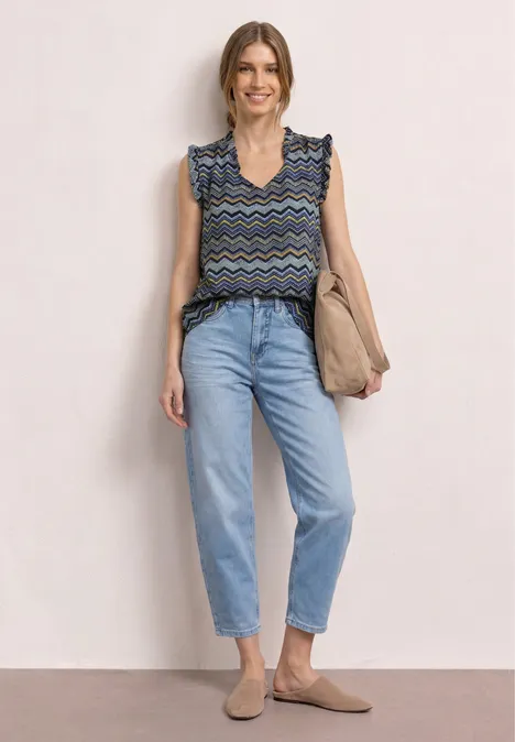 Een vrouw met een gebloemde top met ruches, hoge jeans, beige schoenen en tas voor een neutrale achtergrond.