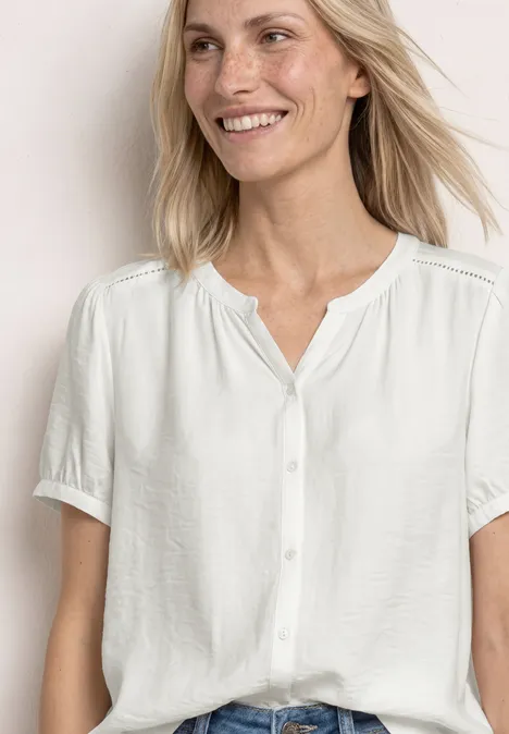Eine Frau mit hellblondem Haar trägt eine weiße, kurzärmelige Bluse mit Knöpfen.