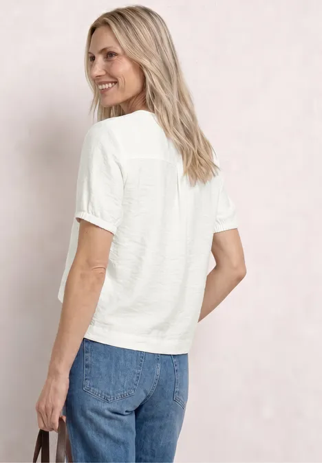 Kurzarm Bluse mit Split Neck und Knöpfen off white Frau von hinten in weißem T-Shirt und blauen Jeans, hält schwarze Tasche, lockere blonde Haare, neutraler Hintergrund.