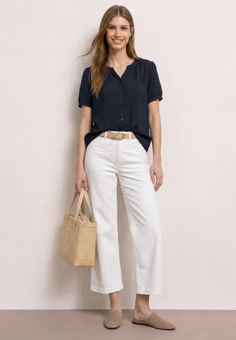 Kurzarm Bluse mit Split Neck und Knöpfen deep blue Frau in dunkelblauer Bluse, weißen Culottes, beigen Loafers und Basttasche vor hellem Hintergrund.