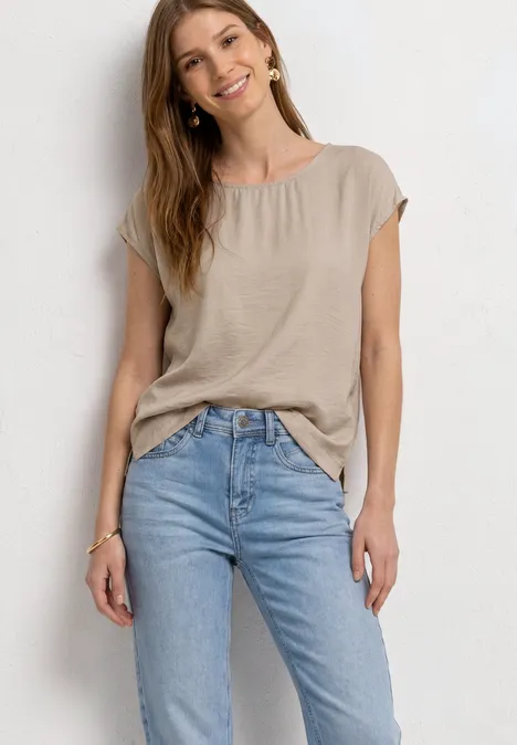 Een vrouw draagt een beige T-shirt met korte mouwen en een lichtblauwe jeans, staand voor een witte achtergrond.
