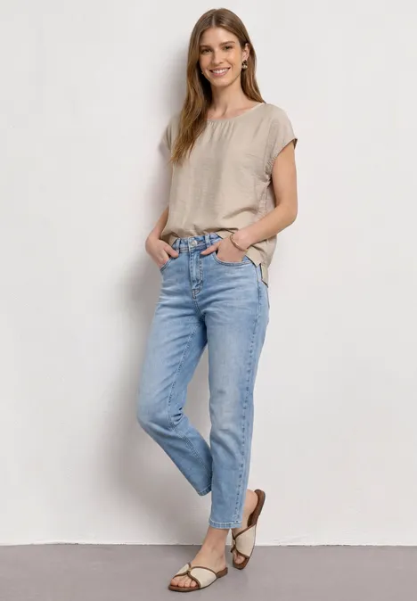 Vrouw in beige T-shirt en lichtblauwe jeans met sandalen, staat ontspannen voor een witte muur.