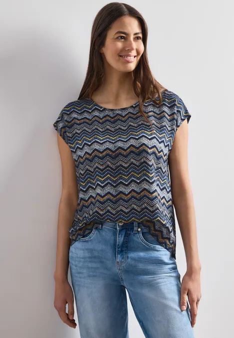 Frau in einem kurzärmligen, gemusterten Oberteil und blauen Jeans vor hellem Hintergrund.