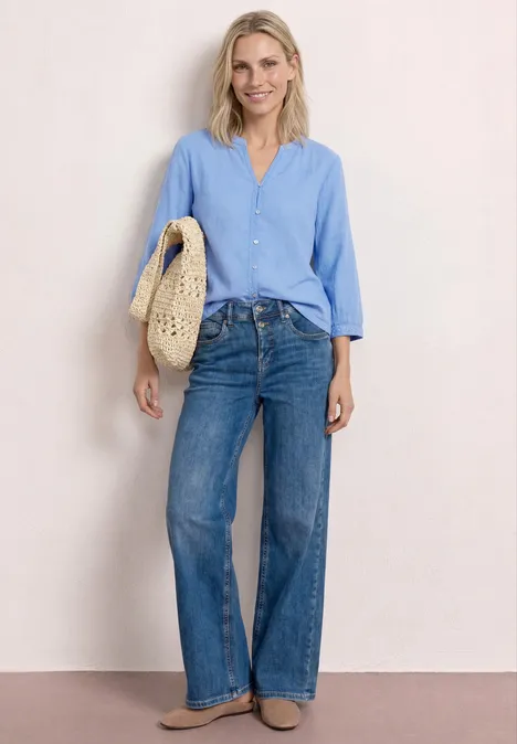 Frau in hellblauer Bluse, weitem Jeans und Tragetasche aus geflochtenem Stroh vor hellem Hintergrund.