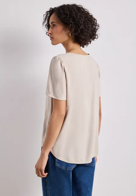 Blouse met ronde hals en korte pofmouwen cotton beige Vrouw van achteren in een beige top met korte mouwen en blauwe spijkerbroek voor een lichte achtergrond.