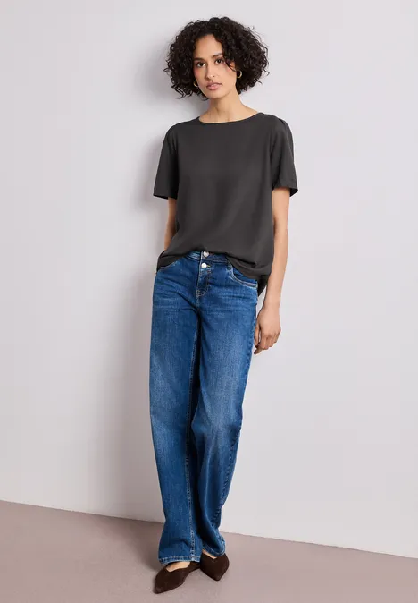 Frau mit lockigem Haar lehnt an weißer Wand und trägt schwarzes T-Shirt, gerade blaue Jeans und braune Loafers.