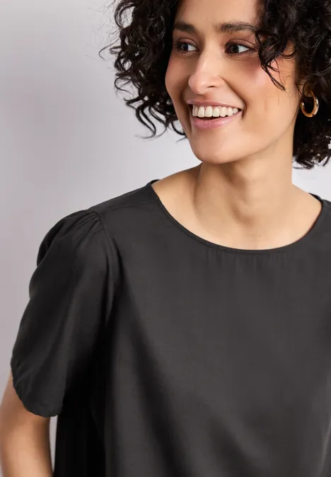 Nahaufnahme einer Person in schwarzer Bluse mit Rundhalsausschnitt, kurzen Ärmeln und goldener Creole.