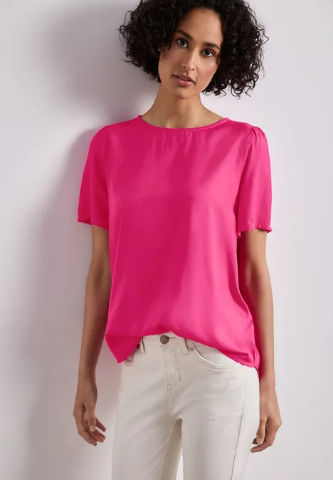 Person mit lockigem Haar trägt pinkes T-Shirt und weiße Hose vor neutralem Hintergrund.