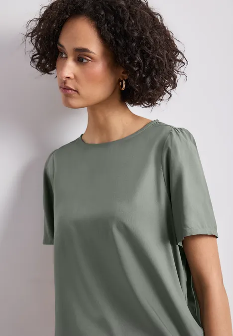 Eine Frau mit lockigem Haar trägt ein schlichtes, olivgrünes Kurzarmshirt vor hellem Hintergrund.