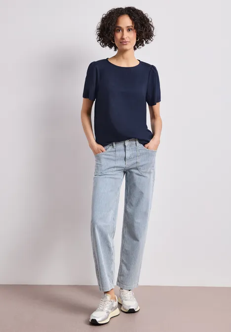 Blouse met ronde hals en korte pofmouwen shadowed navy Een vrouw in een donkerblauw t-shirt, lichte jeans en sneakers staat ontspannen tegen een witte achtergrond.