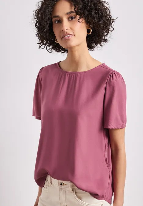 Modell trägt rosa kurzärmlige Bluse mit rundem Ausschnitt und leichtem Fall.