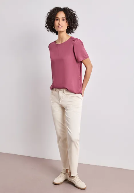 Blouse met ronde hals en korte pofmouwen dark clouded blush Vrouw voor neutrale achtergrond met roze T-shirt, witte broek en witte sneakers.
