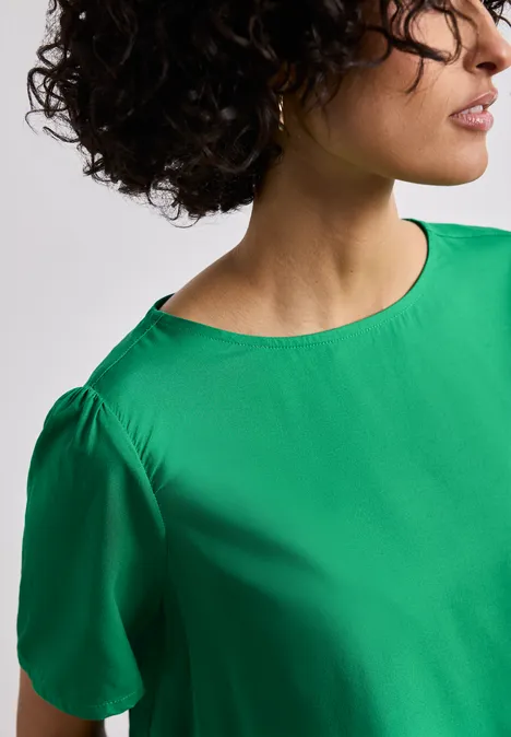 Eine Person trägt eine grüne Bluse mit kurzen Ärmeln, Blick seitlich.