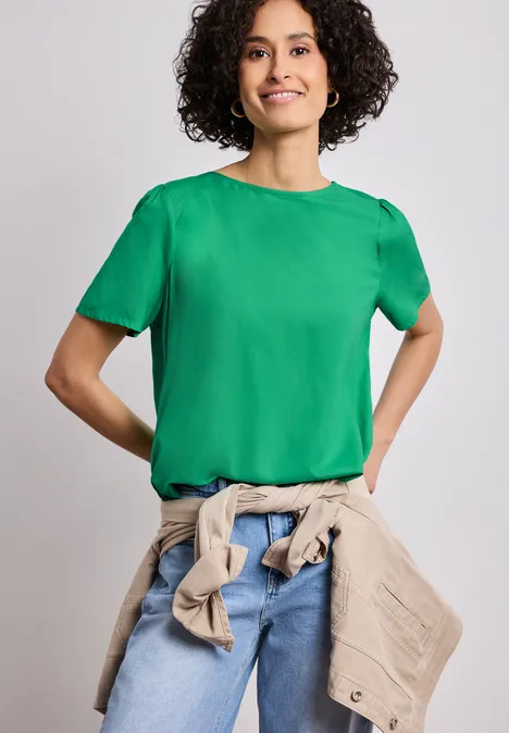 Blouse met ronde hals en korte pofmouwen ever green Persoon met krullend haar draagt een groen T-shirt, spijkerbroek en een beige jas om de taille, voor een neutrale achtergrond.