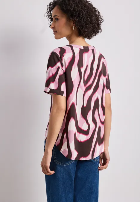 Femme aux cheveux bouclés vue de dos portant un t-shirt rose et noir à motif et un jean bleu.