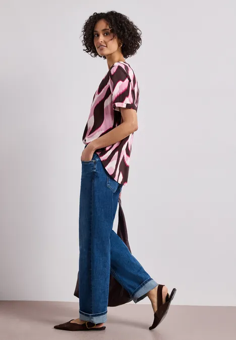 Personne en blouse à motif abstrait avec fente latérale, jean ample et sandales noires, posture de profil.