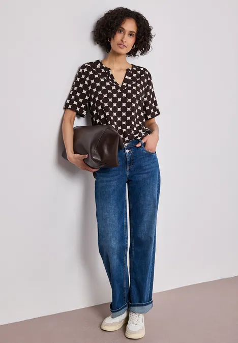 Frau mit lockigem Haar, schwarzer Bluse mit weißen Herzen, Jeans, Sneakern und Tasche lehnt an Wand.