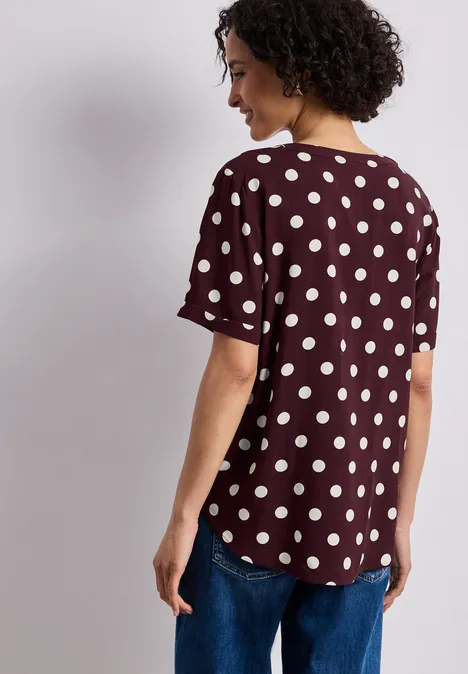Femme de dos portant un t-shirt bordeaux à manches courtes, imprimé à pois blancs, et un jean.