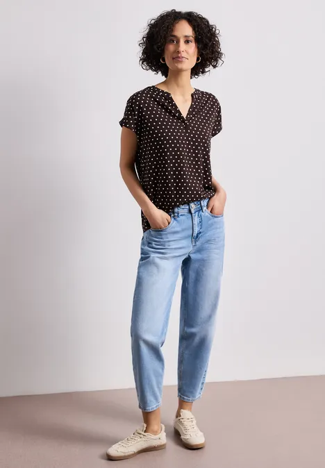 Eine Frau mit kurz geschnittener lockiger Frisur trägt ein schwarzes Polka-Dot-Shirt, blaue Jeans und weiße Sneakers.