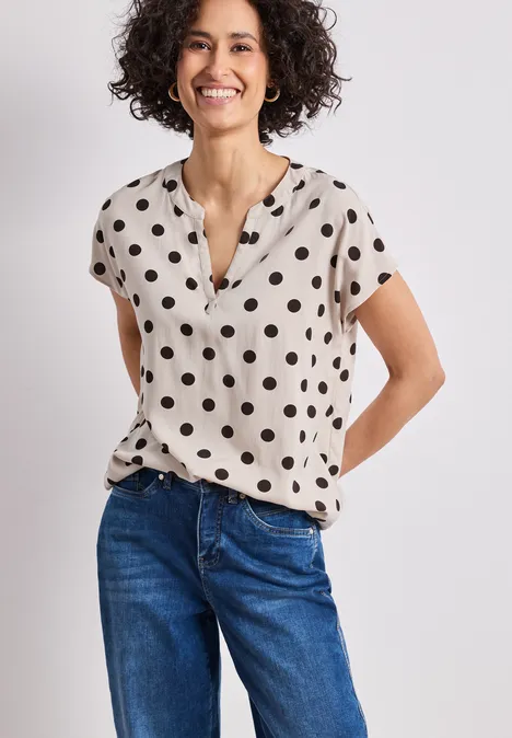 Femme en jean bleu et chemisier beige à pois noirs à manches courtes, mains derrière le dos.