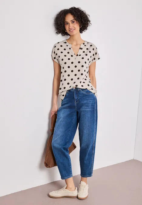 Femme portant un chemisier à pois, un jean bleu et des baskets blanches, adossée à un mur.