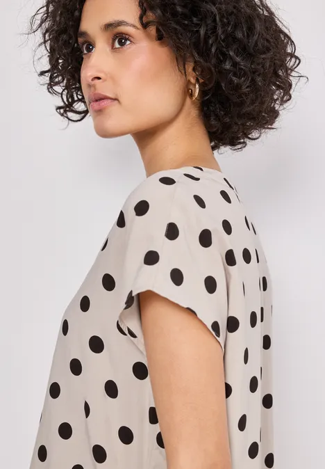 Femme de profil avec cheveux bouclés portant un chemisier beige à pois noirs.