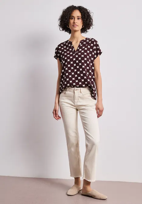 Une femme aux cheveux bouclés porte un chemisier noir à pois blancs, un pantalon clair et des mules.