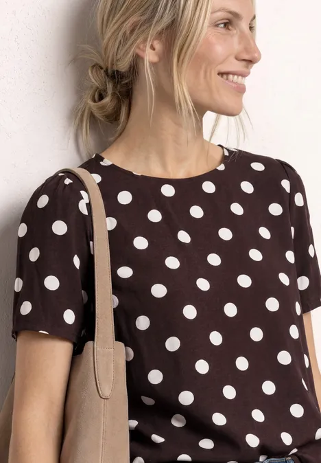 Femme en tee-shirt marron à pois blancs, sac beige à l'épaule, cheveux attachés en chignon lâche.