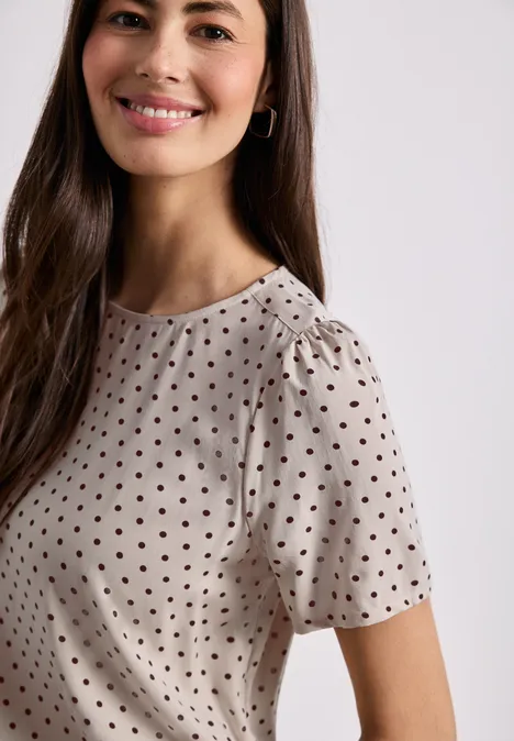 Vrouw draagt een licht topje met zwarte stippen, korte mouwen, ronde hals en subtiele plooitjes op de schouders.