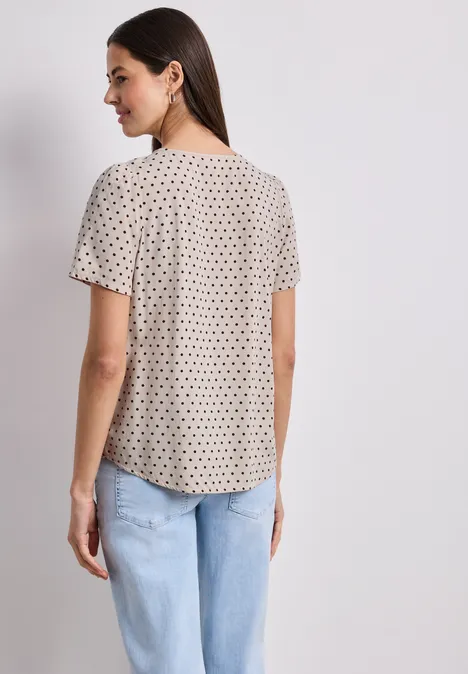 Vrouw van achteren in korte blouse met kleine stippen en lichtblauwe jeans voor een neutrale achtergrond.