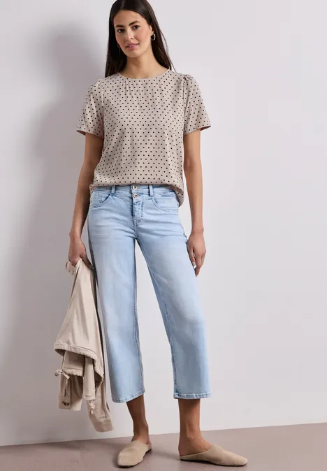 Vrouw in beige T-shirt met print, lichtblauwe cropped jeans, bruine loafers, houdt een beige blazer in de hand.